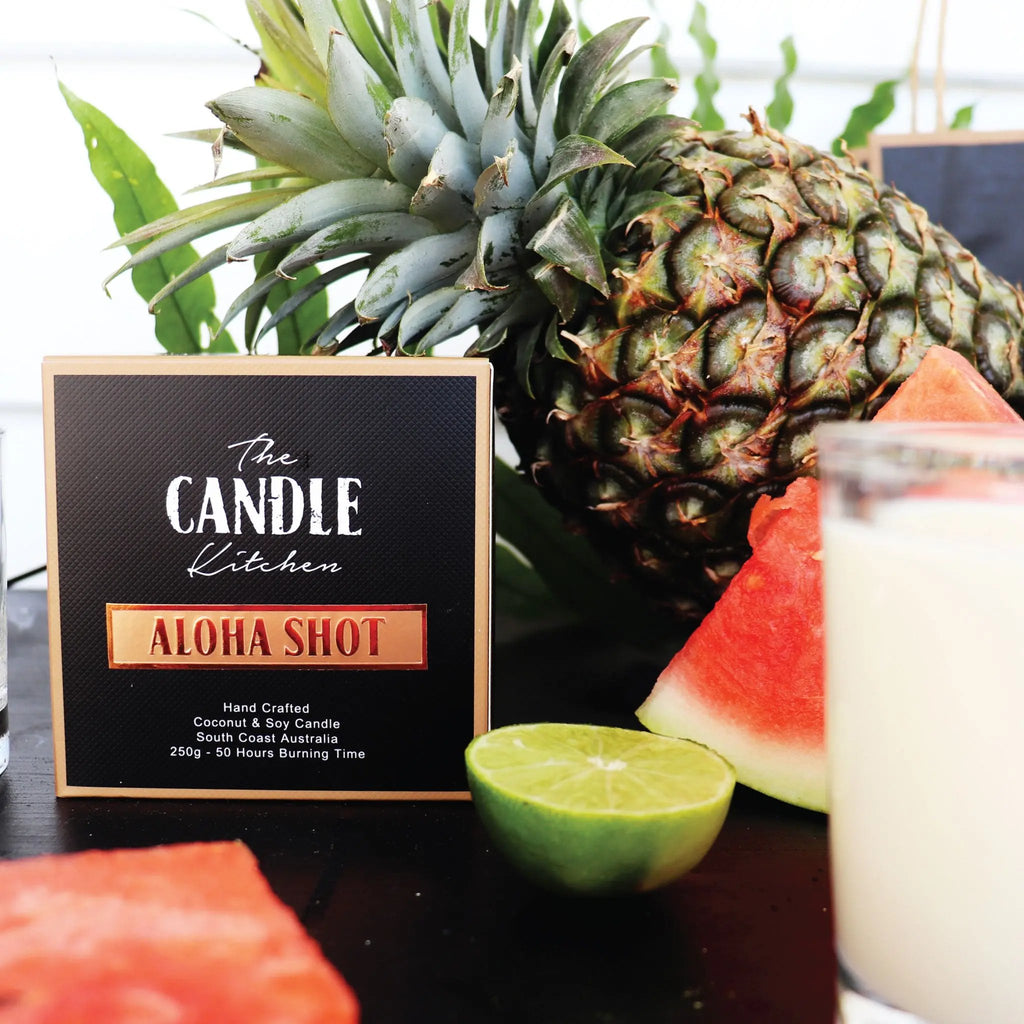 Candle labeled Aloha Shot from the Candle Kitchen with a pineapple, watermelon, and lime on a dark surface.