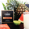 Candle labeled Aloha Shot from the Candle Kitchen with a pineapple, watermelon, and lime on a dark surface.