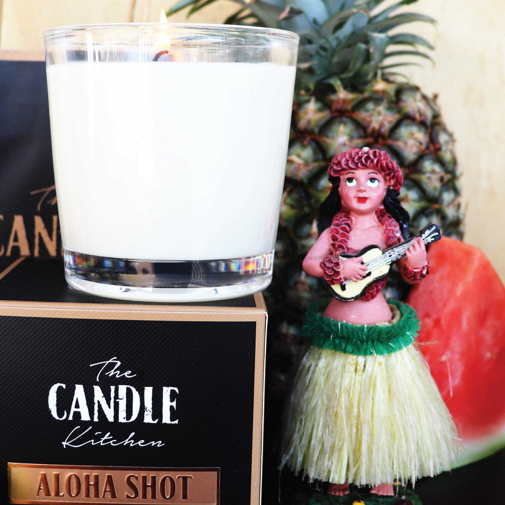 Candle labeled 'Aloha Shot' from The Candle Kitchen with a hula dancer figurine and pineapple in the background.