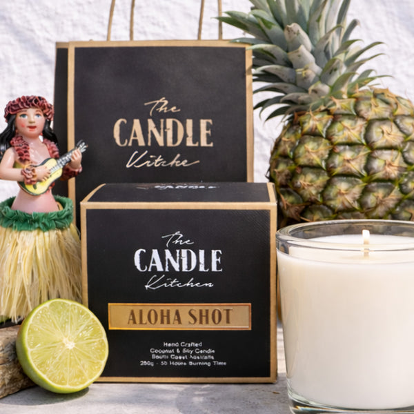 Aloha Shot Soy Candle The Candle kitchen, Candle with 'The Candle Kitchen' packaging, pineapple, hula doll, and lime on a white background