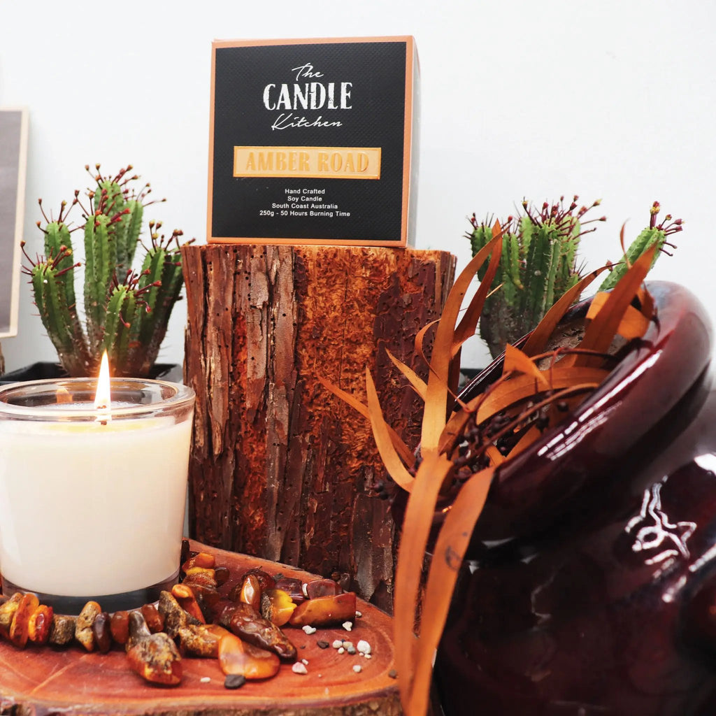 A creamy white Candle with a flickering flame. The Candle Kitchen box standing on a red wood log surrounded by amber stones and a cactus plant in the background.