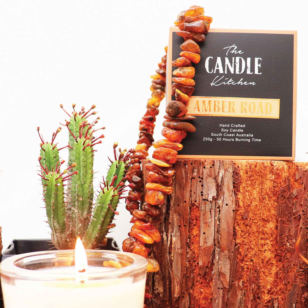 A creamy white Candle with a flickering flame. The Candle Kitchen box standing on a red wood log surrounded by amber stones and a cactus plant in the background.