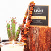 A creamy white Candle with a flickering flame. The Candle Kitchen box standing on a red wood log surrounded by amber stones and a cactus plant in the background.
