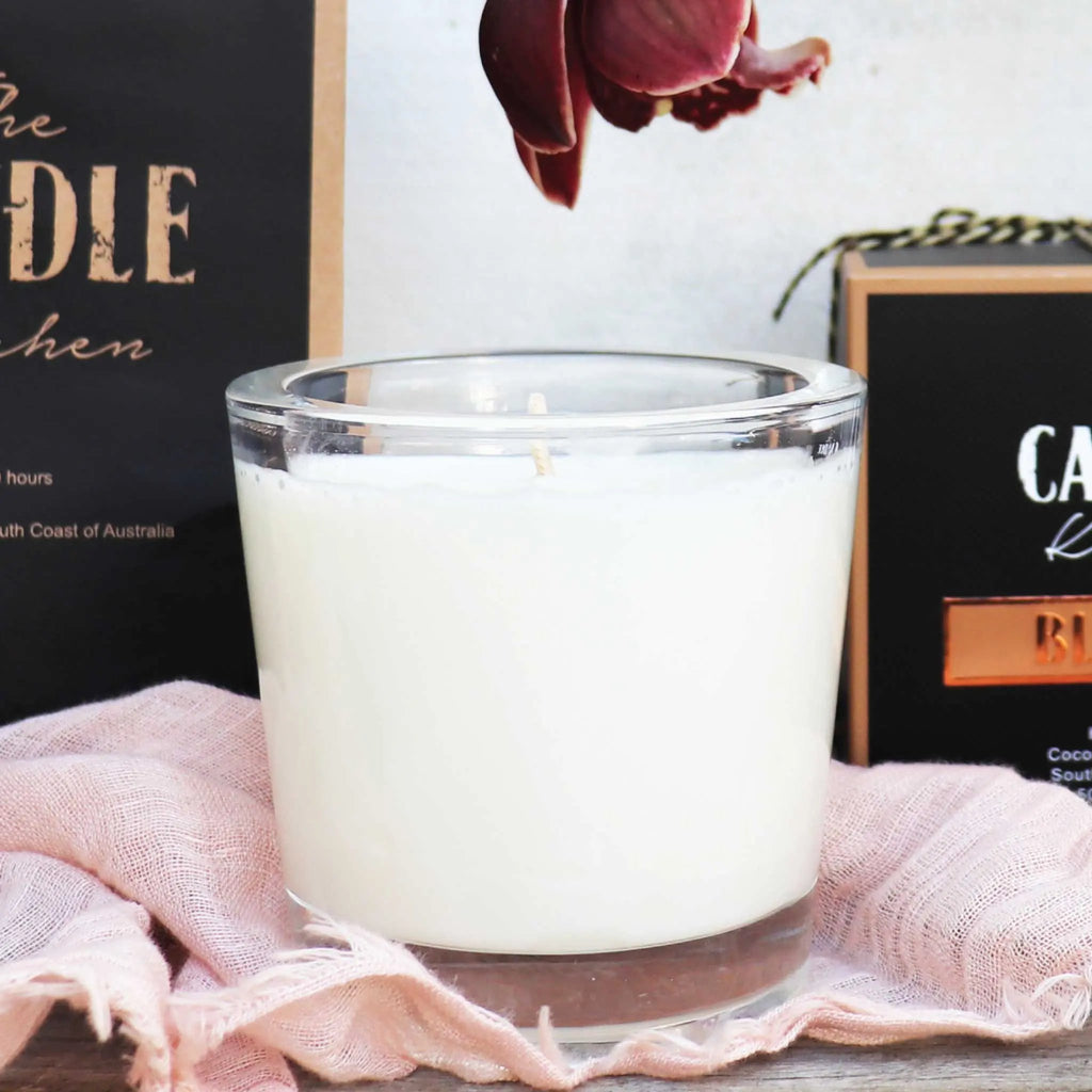 A creamy white candle in a glass jar with decorative boxes and blush linen fabric in the foreground.