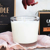 A creamy white candle in a glass jar with decorative boxes and blush linen fabric in the foreground.