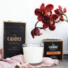 A creamy white candle in a glass jar with decorative boxes and blush linen fabric in the foreground. Surrounded by a Burgundy orchid flower.