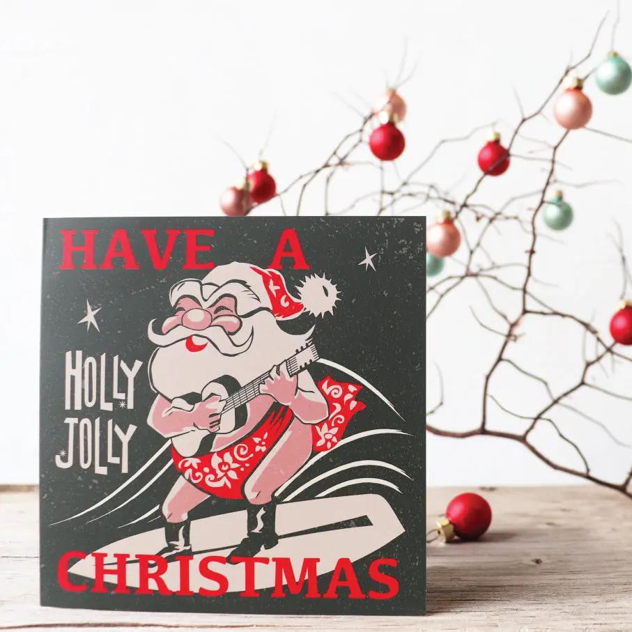 A Christmas card with a graphic of Santa Claus and the text 'HAVE A HOLY JOLLY CHRISTMAS' in red and white on a black background, displayed on a wooden surface with holiday decorations in the background.
