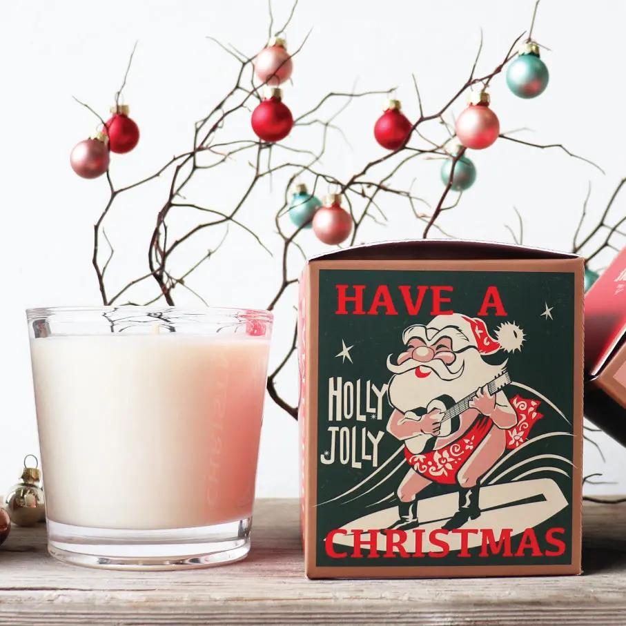 A soy candle with a Christmas-themed label featuring an illustration of Santa Claus and the text 'HAVE A HOLY JOLLY CHRISTMAS'. The candle is placed in front of a glass with Christmas decorations and behind it is a cup of a milk-based drink.