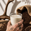 Cuban Coffee soy candle A hand with tattoos holds a clear glass jar candle filled with white wax, styled with coffee beans, a burlap sack, and antlers against a sunlit wall.