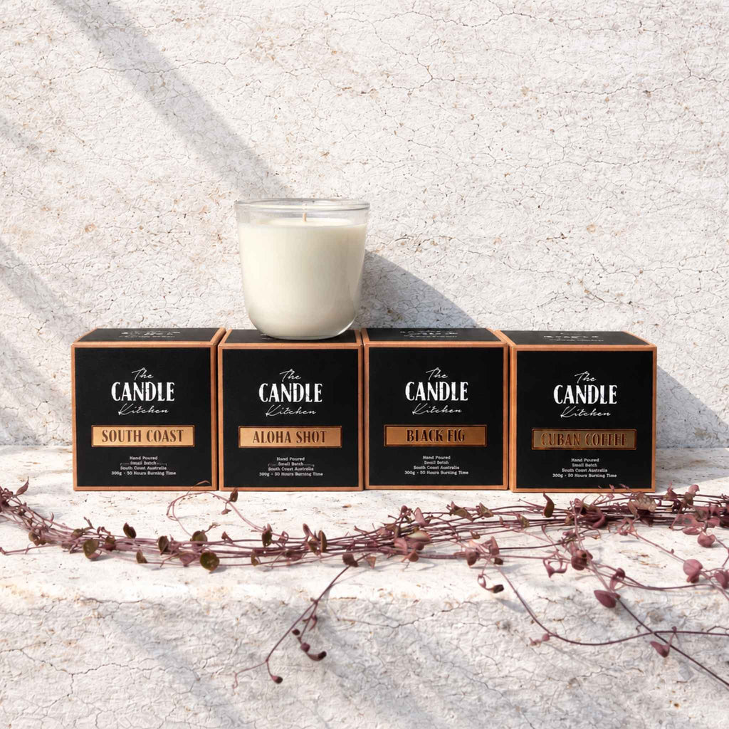 Four black candle boxes with copper trim labeled The Candle Kitchen Collection, arranged on a textured light stone surface with a glass candle in front of them and dried vines in the foreground.
