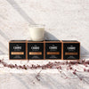 Four black candle boxes with copper trim labeled The Candle Kitchen Collection, arranged on a textured light stone surface with a glass candle in front of them and dried vines in the foreground.