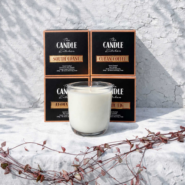 Four black candle boxes with copper-toned borders and white lettering are arranged behind a clear glass jar candle filled with white wax. The labels read South Coast, Cuban Coffee, Aloha shot and Black Fig from the Candle Kitchen