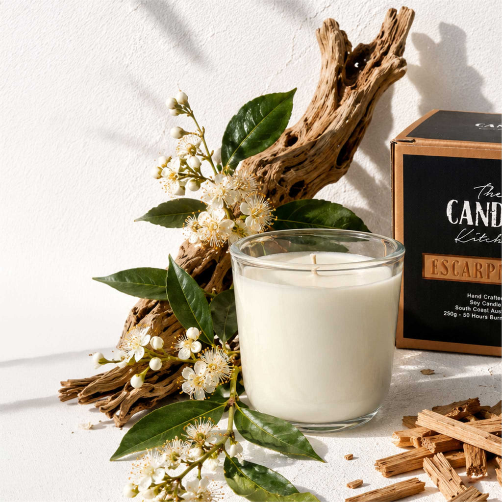 A glass jar candle filled with white wax sits beside its branded box on a white surface, styled with driftwood, green leaves, small white flowers, and wood chips. The box reads 'The CANDLE Kitchen Escarpment and 'Hand Crafted Soy Candle' with 250g, 10 Hours Burn.