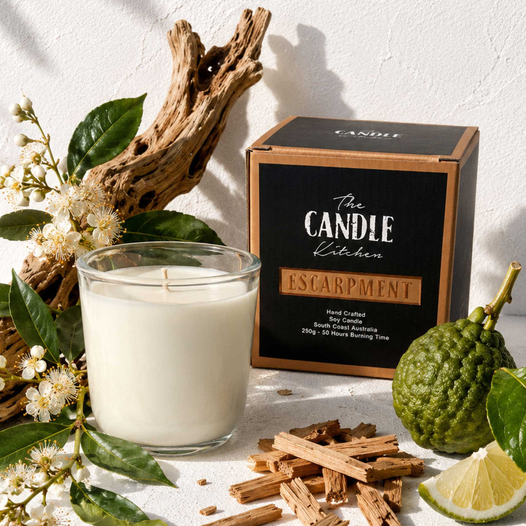 A scented Escarpment soy candle in a clear glass jar sits beside its black and tan branded box on a light textured surface, styled with citrus, cinnamon sticks, flowers, and driftwood. The Candle Kitchen