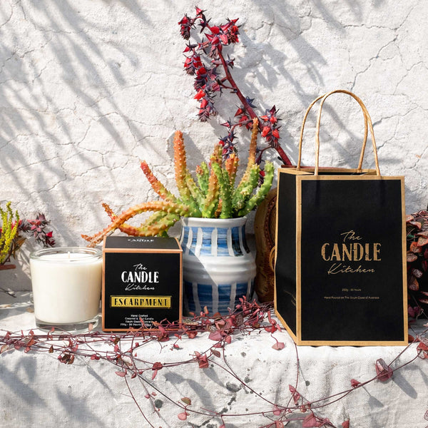 Candle and packaging from 'The Candle Kitchen' on a textured surface with plants and flowers.