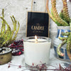 A soy candle named Escarpment, made by The Candle Kitchen, housed in a clear glass jar displayed alongside a box with the product name, set against a rustic backdrop with Succulents and cactuses