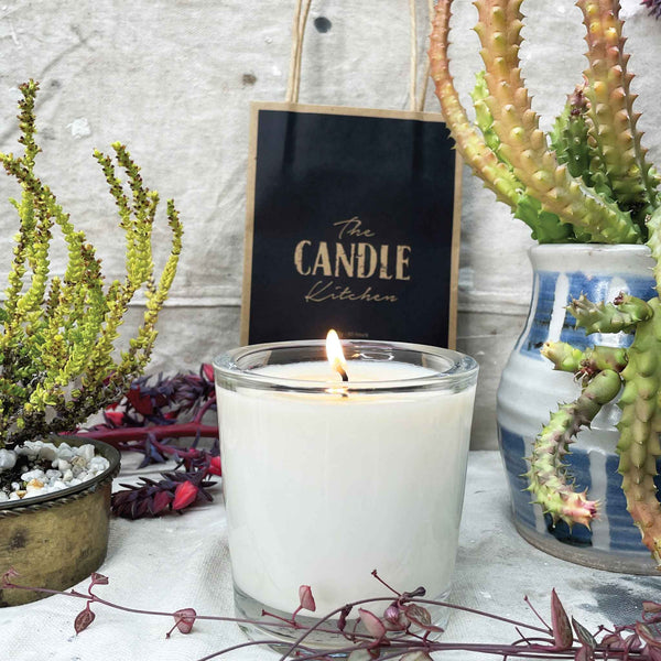 A soy candle named Escarpment, made by The Candle Kitchen, housed in a clear glass jar displayed alongside a box with the product name, set against a rustic backdrop with Succulents and cactuses