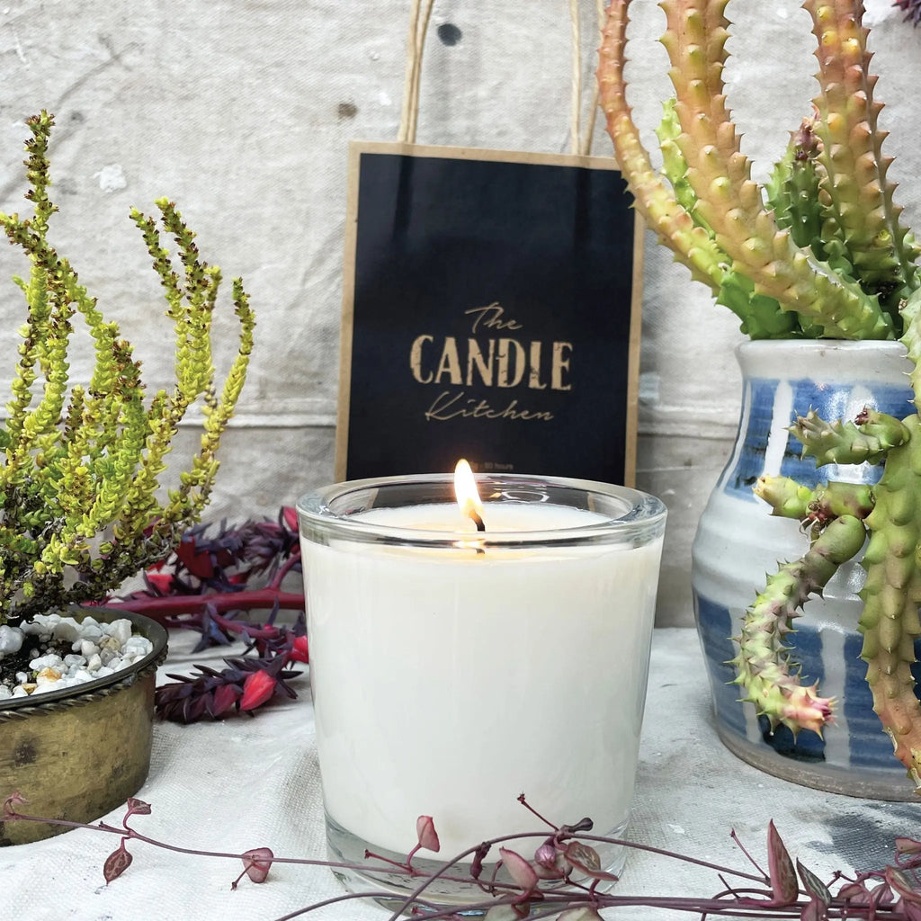 A soy candle named Escarpment, made by The Candle Kitchen, housed in a clear glass jar displayed alongside a box with the product name, set against a rustic backdrop with Succulents and cactuses