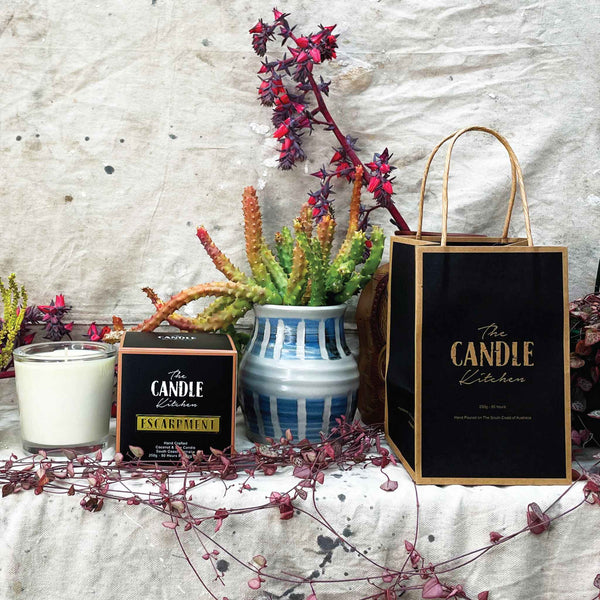 A soy candle named Escarpment, made by The Candle Kitchen, housed in a clear glass jar with a black lid, displayed alongside a box with the product name, set against a rustic backdrop with dried plants.