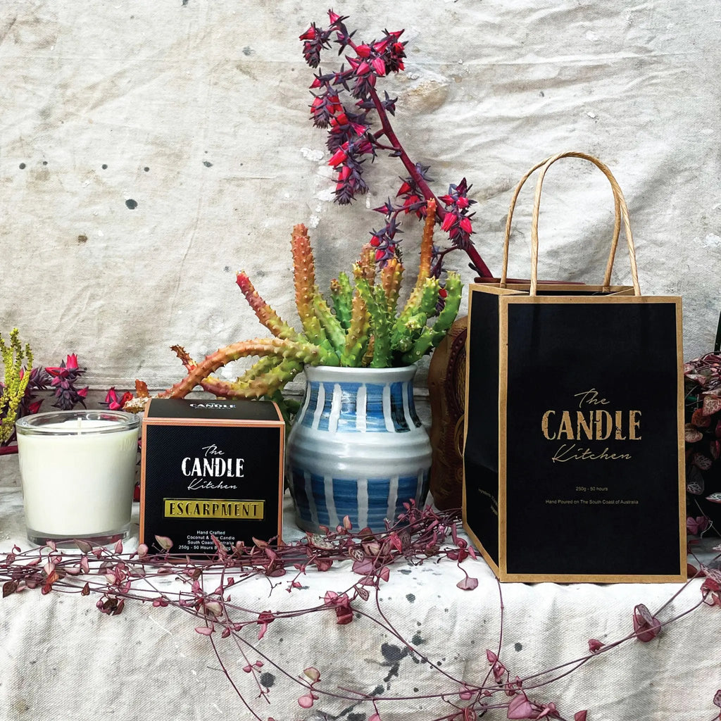 A soy candle named Escarpment, made by The Candle Kitchen, housed in a clear glass jar with a black lid, displayed alongside a box with the product name, set against a rustic backdrop with dried plants.