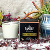 A soy candle named Escarpment, made by The Candle Kitchen, housed in a clear glass jar displayed alongside a box with the product name, set against a rustic backdrop with Succulents and cactuses