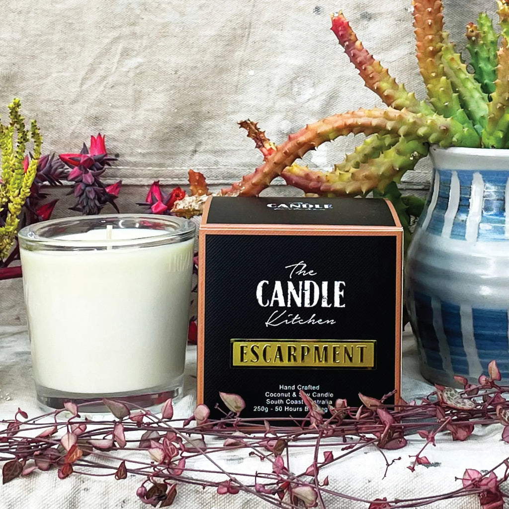 A soy candle next to a black box from the Candle Kitchen, housed in a clear glass jar displayed against a rustic backdrop with Succulents and cactuses. 