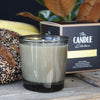 Candle in a glass container with a box labeled The Candle Kitchen on a wooden surface. A seeded loaf of bread behind the candle.