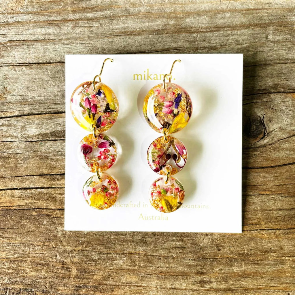 Colorful floral earrings on a white card with 'mikano' branding, placed on a wooden surface.
