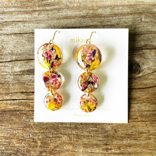 Colorful floral earrings on a white card with 'mikano' branding, placed on a wooden surface.