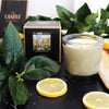 Lemoncello candle with packaging from the Candle Kitchen and lemons on a dark surface. Wet rich green lemon tree leaves in the fore ground.