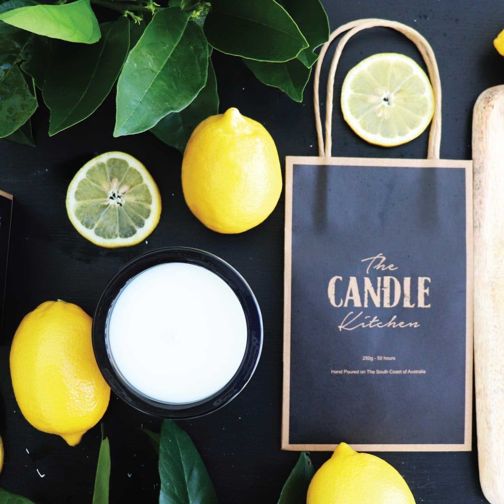 looking down at the top circle of a Candle with bright yellow lemons and 'The Candle Kitchen' bag on a dark surface.