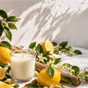 A white candle in a clear glass jar sits on a white draped surface surrounded by lemons, green leaves, small white flowers,  in bright natural light. Lemoncello Soy Candle from the Candle Kitchen