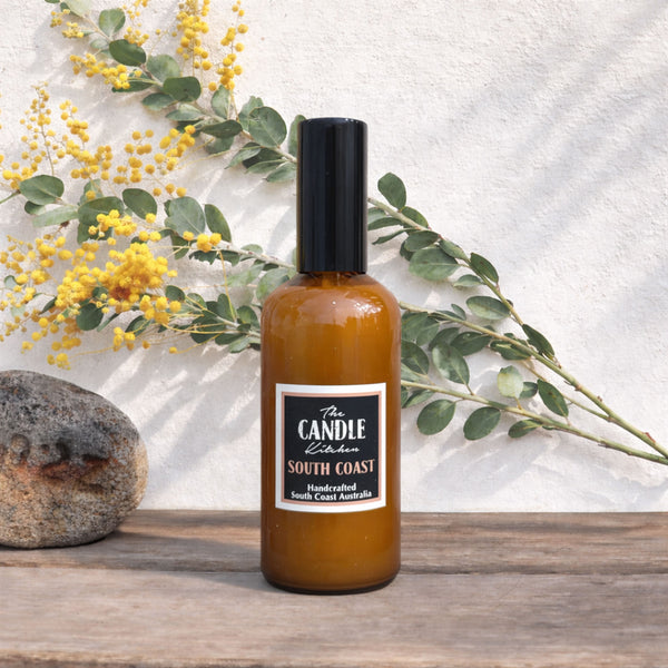 Bottle of The Candle Kitchen South Coast product on a wooden surface with greenery and flowers in the background.