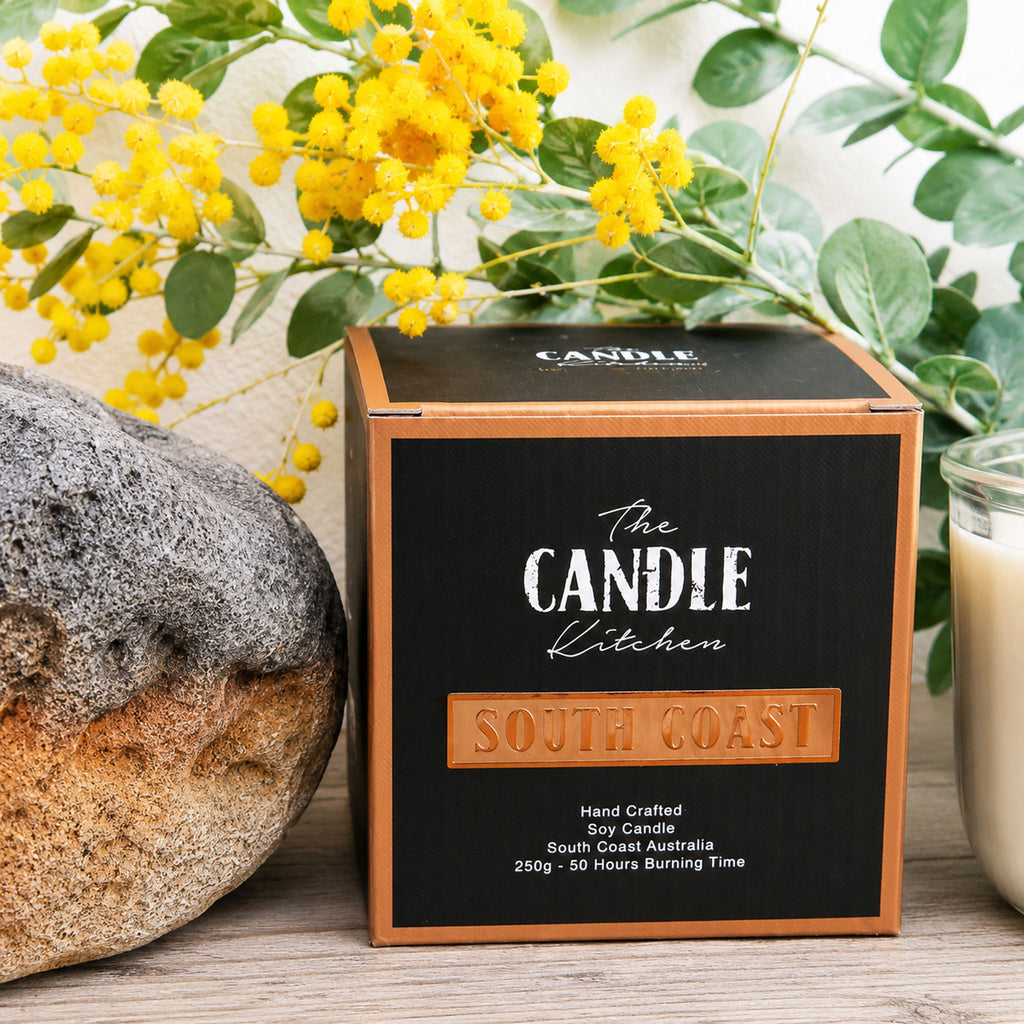 The Candle Kitchen packaging from 'The Candle Kitchen' with yellow wattle flowers and a convict brick in the background