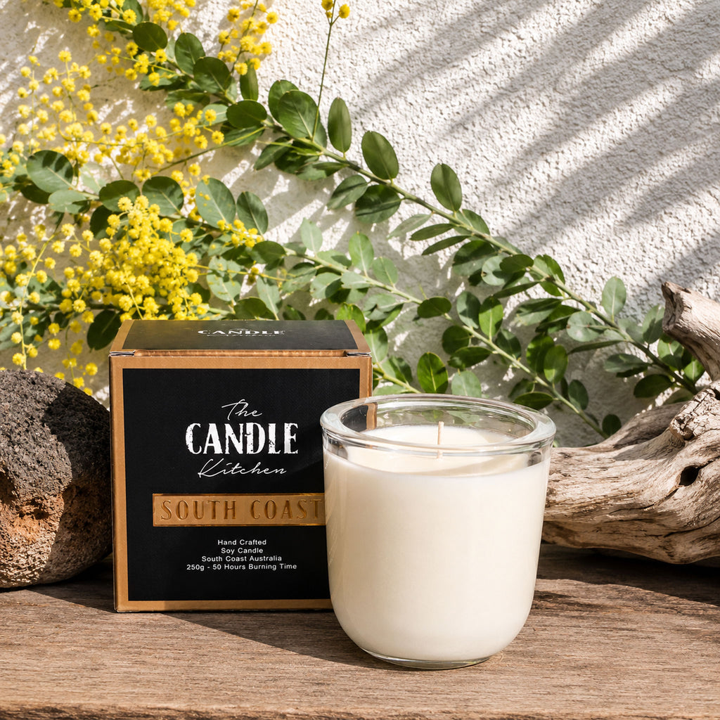 South Coast Soy Candle The Candle kitchen, Candle in a glass holder with a 'The Candle Kitchen' box and yellow flowers on a wooden surface.
