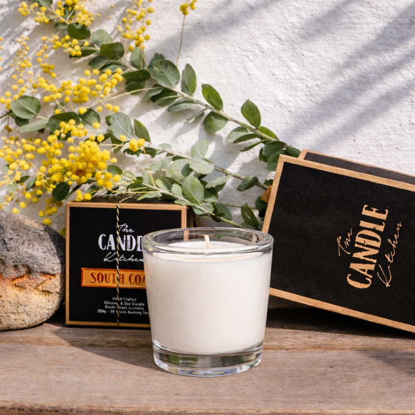 South Coast Soy Candle The Candle kitchen, Candle in a glass holder with a 'The Candle Kitchen' box and yellow flowers on a wooden surface.