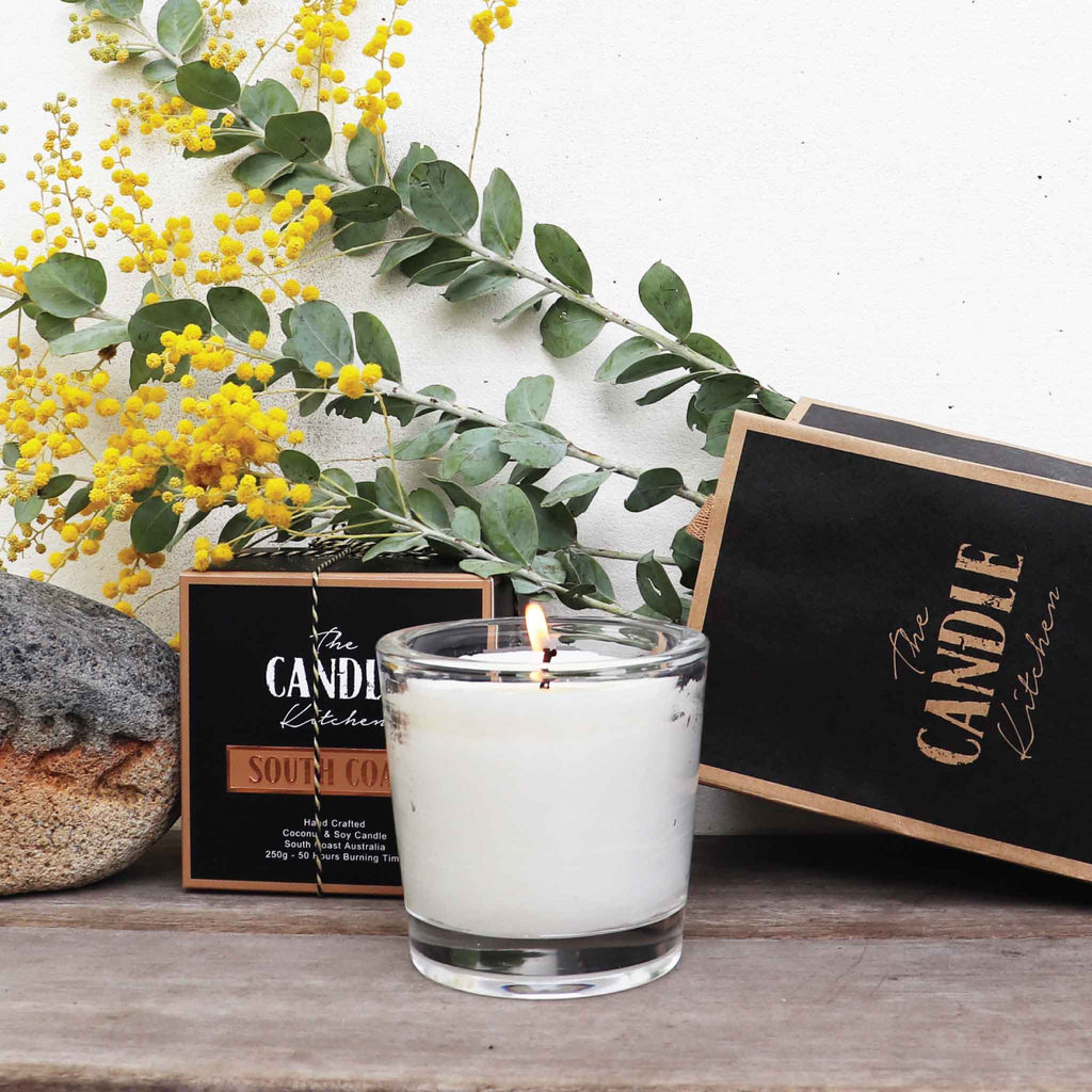 A Creamy white Candle with a flickering flame in front of the Candle Kitchen bag and box with a bunch of wattle in the background.