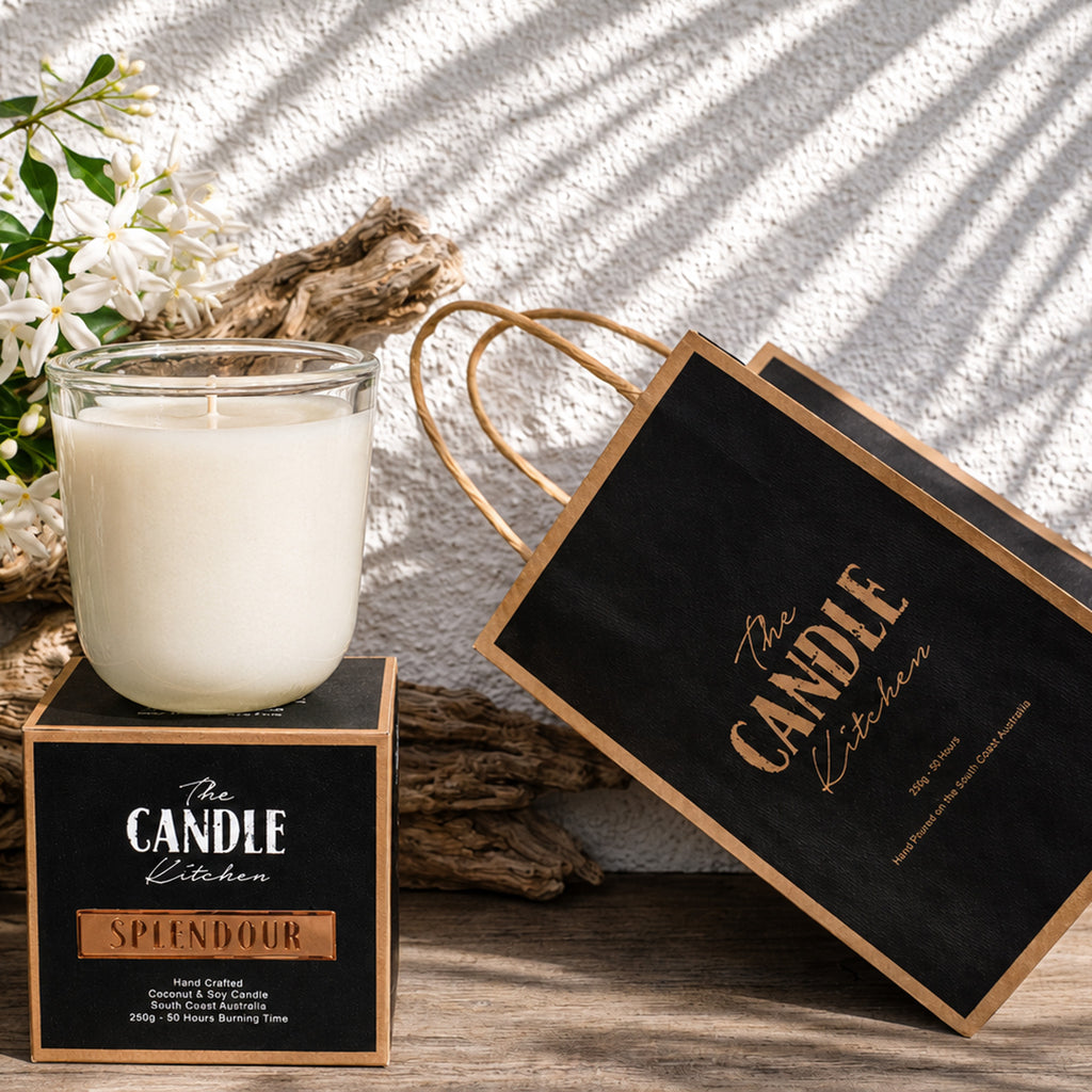 Splendour Soy Candle The Candle kitchen, Candle with packaging and flowers on a textured surface Candle on a stand with a decorative bag labeled 'The Candle Kitchen' on a wooden surface.