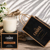 Splendour Soy Candle The Candle kitchen, Candle with packaging and flowers on a textured surface Candle on a stand with a decorative bag labeled 'The Candle Kitchen' on a wooden surface.