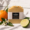 A rectangular bar of handmade soap labeled 'The Candle Kitchen Citrus Grove' sits on a white wood surface, styled with a whole orange, a lime half, green leaves, and a white citrus blossom. The soap is tan/beige with dried orange flower petals on top.