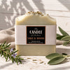 A handmade bar candle wrapped in a cream paper band labeled 'The Candle Kitchen Sage & Wood', styled on a white wooden surface with sage leaves, rosemary, and a beige cloth.